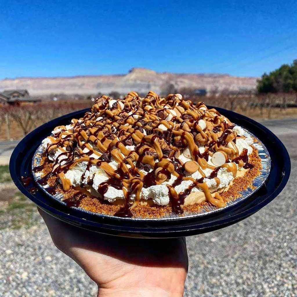 A hand holding a whole cream pie topped with whipped cream, chocolate and caramel drizzle, almonds and chocolate chips, in front of bare orchard rows and the mesa above Palisade.